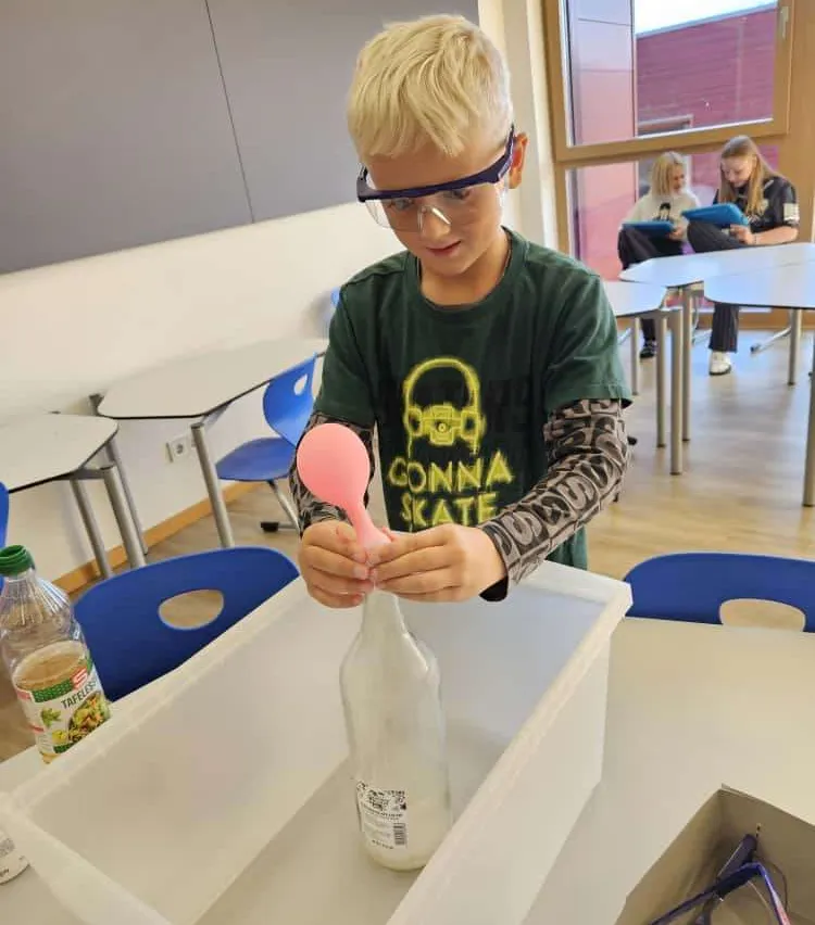 Ein junger Junge trägt Schutzbrillen und hält einen rosa Luftballon, er ist dabei, ihn in eine Flasche in einem Wissenschaftsexperiment freizulassen.