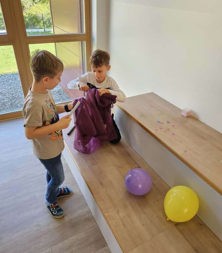 Zwei junge Jungen spielen mit einer lila Tasche in der Nähe einer Bank mit zwei Luftballons, einem lila und einem gelben. Sie befinden sich in einem Raum mit einem Glasfenster, das einen Außenbereich zeigt.