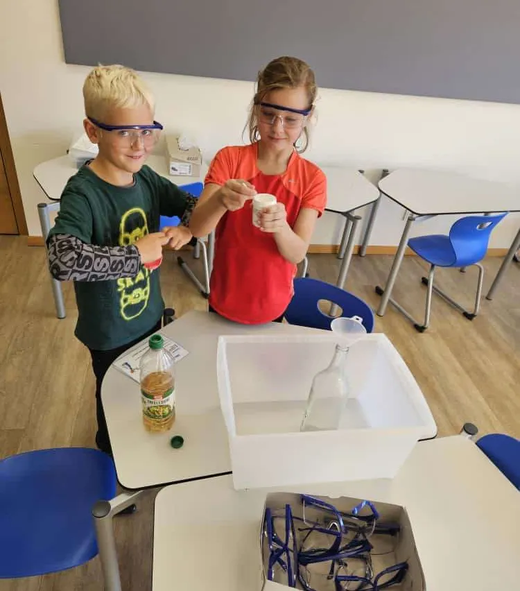 Zwei Kinder führen in einem Klassenzimmer ein wissenschaftliches Experiment durch, tragen Schutzbrillen und halten einen kleinen Behälter. Eine Flasche und ein durchsichtiger Behälter stehen auf dem Tisch vor ihnen.