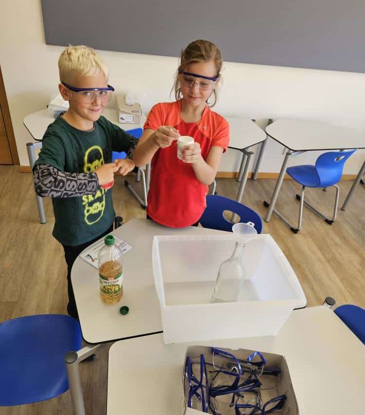 Zwei Kinder führen in einem Klassenzimmer ein wissenschaftliches Experiment durch, tragen Schutzbrillen und halten einen kleinen Behälter. Eine Flasche und ein durchsichtiger Behälter stehen auf dem Tisch vor ihnen.