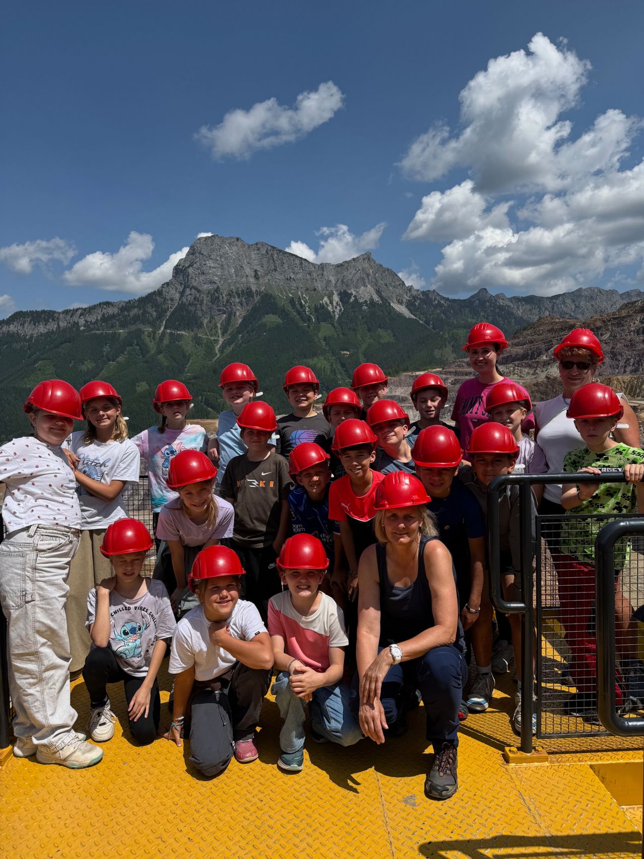 Eine Gruppe von Kindern und Erwachsenen trägt rote Helme, lächelt und posiert für ein Foto mit Bergen im Hintergrund.