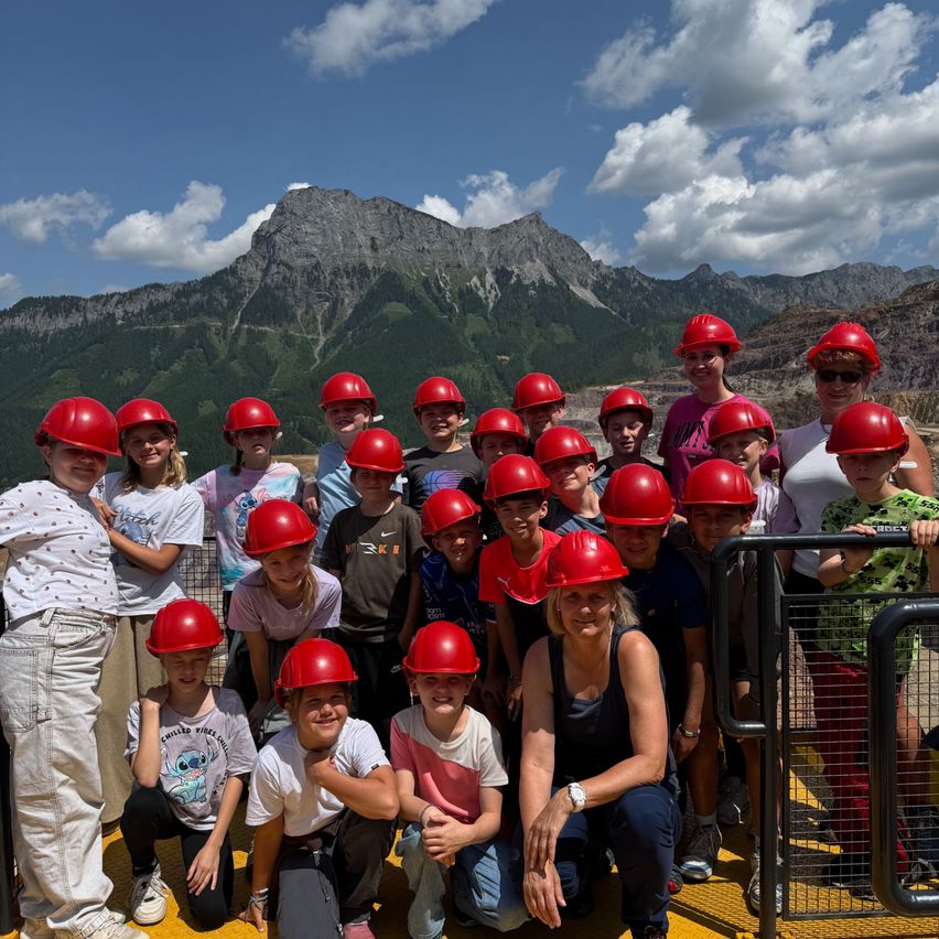 Eine Gruppe von Kindern und Erwachsenen trägt rote Helme, lächelt und posiert für ein Foto mit Bergen im Hintergrund.