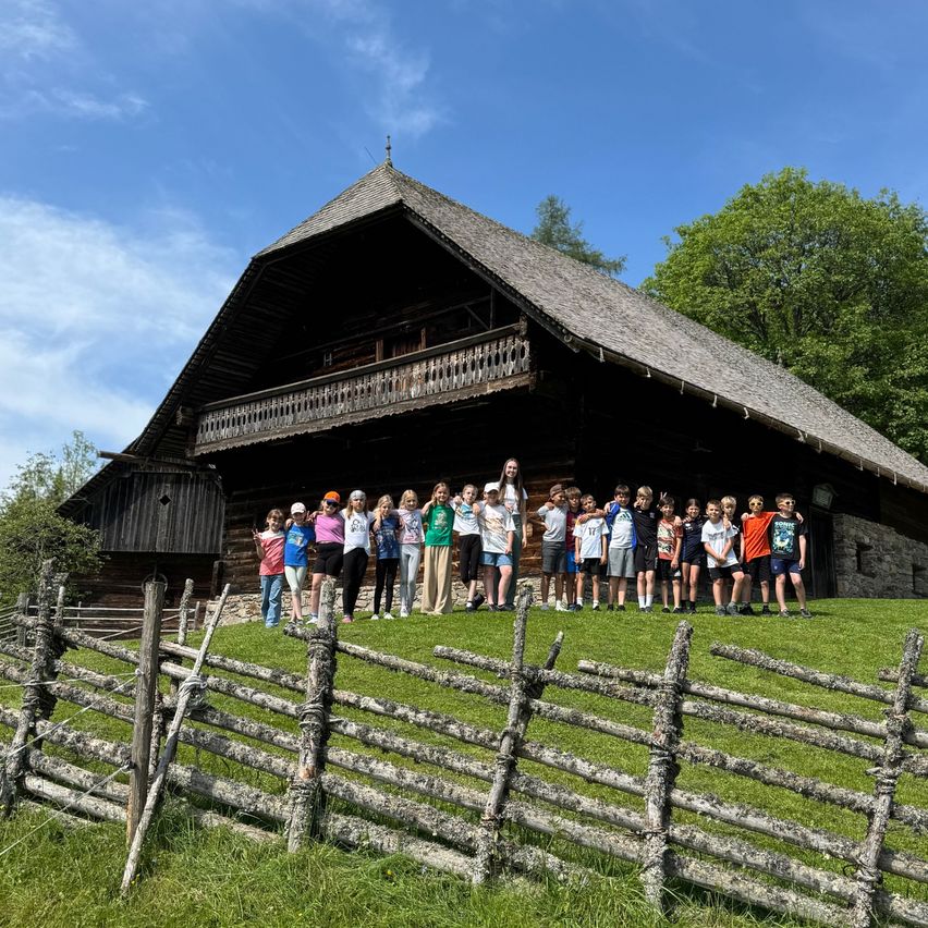 Eine Gruppe von Menschen, wahrscheinlich Schüler, steht vor einem traditionellen Holzhaus mit Steinfundament. Sie posieren für ein Foto an einem sonnigen Tag.