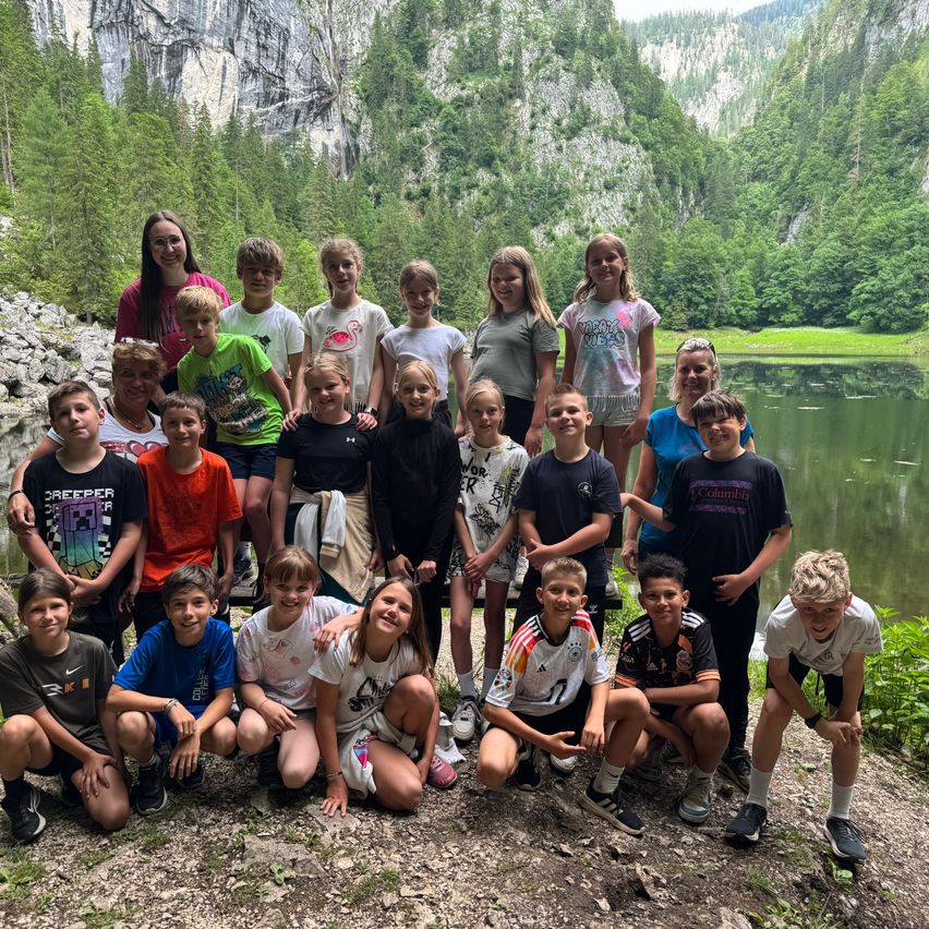 Eine Gruppe von Kindern und Jugendlichen posiert für ein Foto am See, umgeben von Grünflächen und Bergen.