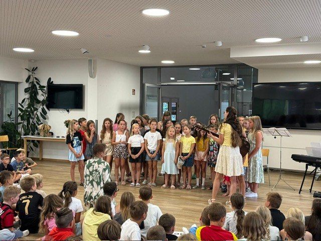 Eine Gruppe von Kindern auf der Bühne, die ein Lied singen, umgeben von einem Publikum aus Kindern, die auf dem Boden sitzen.