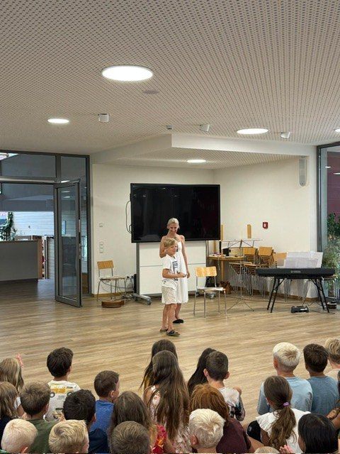 Zwei Mädchen stehen auf einer Bühne in einem Raum mit Klavier und Fernseher. Eine Menschenmenge von Kindern sitzt auf dem Boden und schaut zu.