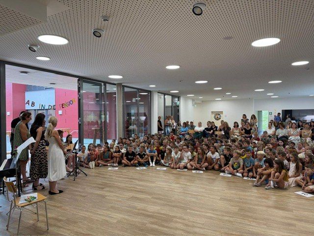Eine Gruppe von Kindern und Erwachsenen sitzt auf einem Holzboden in einem Raum mit Glaswänden. Eine Frau steht vor ihnen, möglicherweise eine Lehrerin. Ein Klavier und ein Notenständer sind in der Nähe. Der Raum hat eine Decke mit Lichtern und einem Ausfahrtsschild.