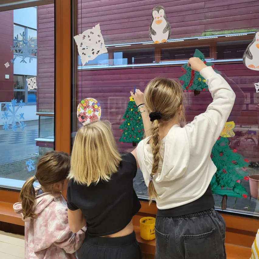 Drei Frauen schmücken ein Fenster mit Weihnachtsbäumen und Schneeflocken. Die Frau in der Mitte hält einen blauen Stift.