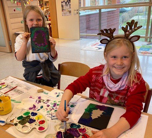 Zwei junge Mädchen in einem Klassenzimmer, eine mit Rentierhörnern, malt auf einer Leinwand, während die andere ein fertiges Werk in der Hand hält.