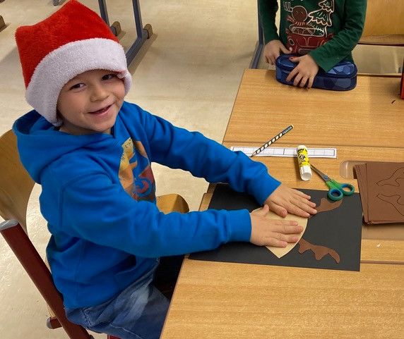 Ein junger Junge in einer blauen Kapuzenjacke und einer Weihnachtsmütze lächelt, während er an seinem Schreibtisch ein Bastelprojekt ausführt.