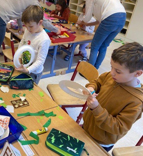 Mehrere Kinder sind in einem Klassenzimmer mit einer Kunstaktivität beschäftigt. Ein Junge bastelt einen Papierkreis, während ein anderer einen Papierkreis ausschneidet. Hinter ihnen steht eine Frau, möglicherweise eine Lehrerin. Verschiedene Bastelmaterialien sind auf den Tischen verstreut.