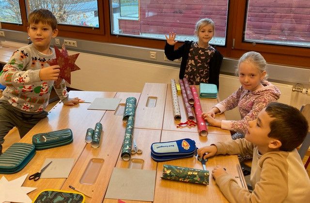 Vier Kinder sitzen um einen Tisch mit Bastelmaterial versammelt, wahrscheinlich in einem Klassenzimmer. Ein Junge hält einen Stern, während ein Mädchen lächelt und mit der Hand zeigt. Auf dem Tisch liegen Scheren, Geschenkpapier und ein Stiftetui.