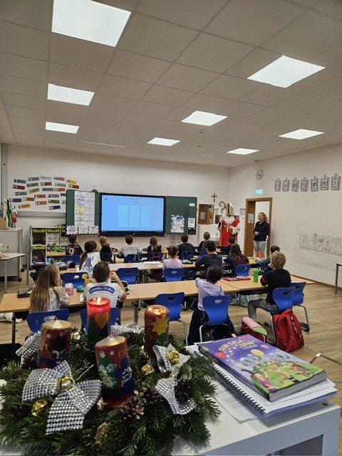 Ein Klassenzimmer mit Schülern an Tischen, einem Lehrer in Santa-Kleidung neben einer Leinwand und einer Lehrerin neben einer Tür. Kerzen und Bücher sind auf den Tischen.