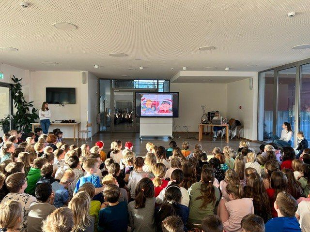 Ein Klassenzimmer voller Kinder, einige tragen Mützen, sitzen auf dem Boden. Eine Frau steht vor einem Bildschirm, der einen Cartoon zeigt. Eine andere Frau sitzt an einem Schreibtisch mit Laptop.