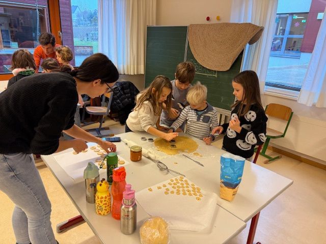 Eine Gruppe von Kindern und einem Erwachsenen arbeitet gemeinsam in einem Klassenzimmer. Sie backen Kekse auf einem Tisch mit verschiedenen Zutaten und Utensilien.