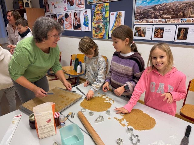 Eine ältere Frau bäckt mit einer Gruppe von Kindern in einem Klassenzimmer Kekse. Verschiedene Backutensilien liegen auf dem Tisch.