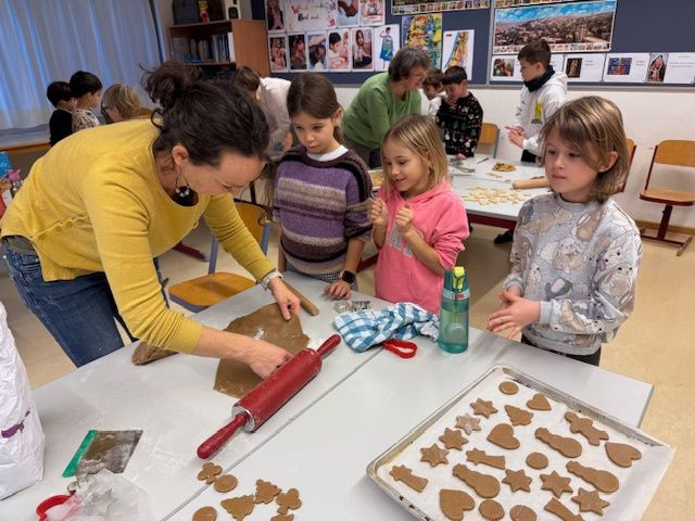 Eine Gruppe von Kindern und Erwachsenen versammelt sich um einen Tisch und stellt Lebkuchenplätzchen her. Eine Frau in einem gelben Shirt rollt Teig mit einem roten Nudelholz aus.