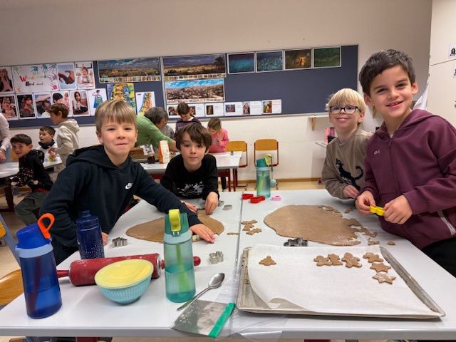 Fünf Kinder backen in einem Klassenzimmer Kekse, umgeben von Ausstechformen, einer Ablage und einer Schüssel auf dem Tisch. Ein Kind benutzt eine Keksausstecher, während andere lächeln und in die Kamera schauen. Es gibt Poster an der Wand und andere Kinder und Lehrer im Hintergrund.