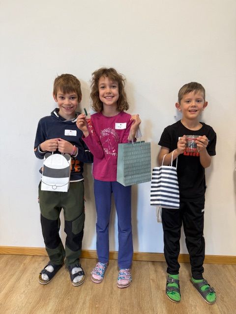 Drei Kinder stehen zusammen auf einem Holzboden, lächeln und halten Papiertüten. Das Mädchen in der Mitte trägt ein rosa T-Shirt und blaue Hose. Der Junge auf der linken Seite trägt einen blauen Hoodie und grüne Hose. Der Junge auf der rechten Seite trägt ein schwarzes T-Shirt und schwarze Hose. Sie tragen alle Sneakers.