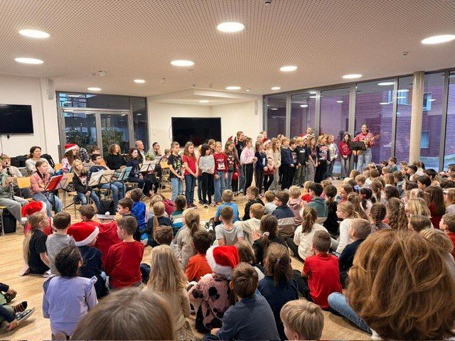 Eine Gruppe von Kindern und Erwachsenen versammelt sich in einem Raum, wahrscheinlich für eine musikalische Aufführung. Sie tragen Weihnachtsmützen. Einige von ihnen halten Musikinstrumente. Der Raum hat einen Holzboden und Glaswände.