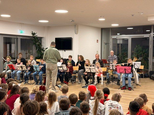 Eine Gruppe von Kindern und Erwachsenen spielt Musikinstrumente auf der Bühne. Sie sind in Feiertagskleidung gekleidet und von einem Publikum umgeben.