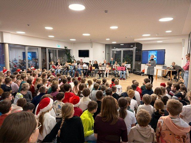 Eine Gruppe von Kindern und Erwachsenen in einem Raum mit einer großen Leinwand und Musikständen, einige tragen Weihnachtsmützen. Eine Frau steht mit einem Mikrofon auf der Bühne.
