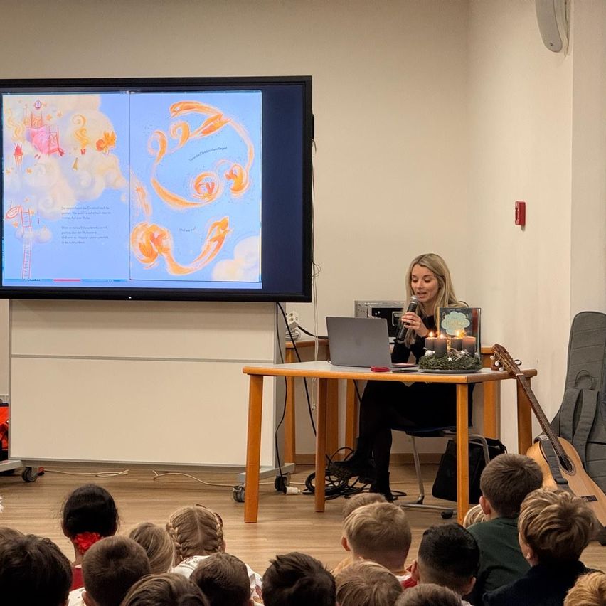 Eine Frau liest vor einem Tisch in einem Klassenzimmer aus einem Buch, während ein großer Bildschirm ein Bild hinter ihr zeigt. Mehrere Kinder beobachten aufmerksam.