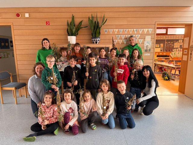 Gruppenfoto von Kindern und Erwachsenen, die Pflanzenzweige halten und in einem Klassenzimmer mit Holzwänden und einem Banner mit der Aufschrift 'Willkommen' lächeln.
