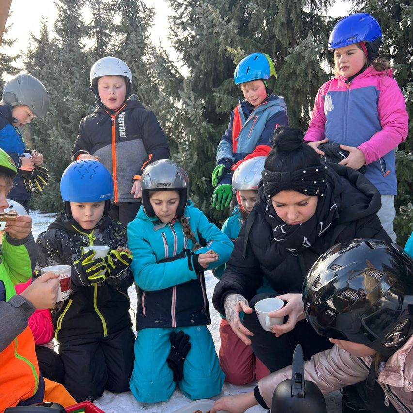 Eine Gruppe von Kindern und Erwachsenen sitzt in einem verschneiten Bereich. Sie tragen alle Winterjacken und Helme. Sie haben wahrscheinlich einen Snack. Die erwachsene Frau in der Mitte hält eine Tasse und scheint mit den Kindern zu sprechen. Hinter ihnen stehen Tannenbäume.