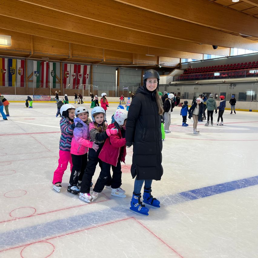 Eine Frau steht auf einer Eisbahn mit vier Kindern, alle in Winterkleidung und Helmen. Sie stehen in der Nähe des Randes der Eisbahn, mit anderen Skatern im Hintergrund.