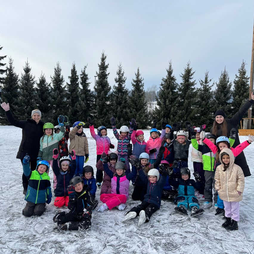 Eine Gruppe von Kindern und Erwachsenen posiert für ein Foto auf einem verschneiten Feld mit Tannen im Hintergrund. Sie tragen alle Winterkleidung und einige haben Helme auf.