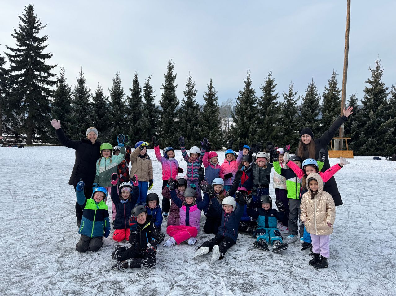 Eine Gruppe von Kindern und Erwachsenen posiert für ein Foto auf einem verschneiten Feld mit Tannen im Hintergrund. Sie tragen alle Winterkleidung und einige haben Helme auf.