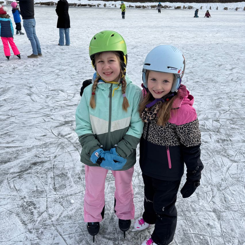 Zwei junge Mädchen, beide mit Helmen und Eislaufschuhen, posieren für ein Foto auf einem zugefrorenen See. Hinter ihnen stehen oder skaten mehrere andere Personen.