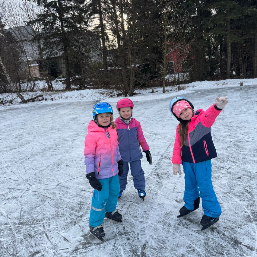 Drei junge Mädchen eislaufen auf einer Eisbahn im Freien. Sie tragen Winterkleidung und Helme. Schnee und Bäume umgeben die Eisbahn.
