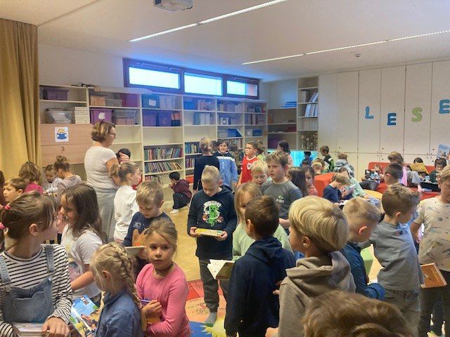 Eine Gruppe von Kindern, wahrscheinlich in einer Bibliothek, sind versammelt, halten Bücher in den Händen und lauschen einer Frau, die vor ihnen steht.