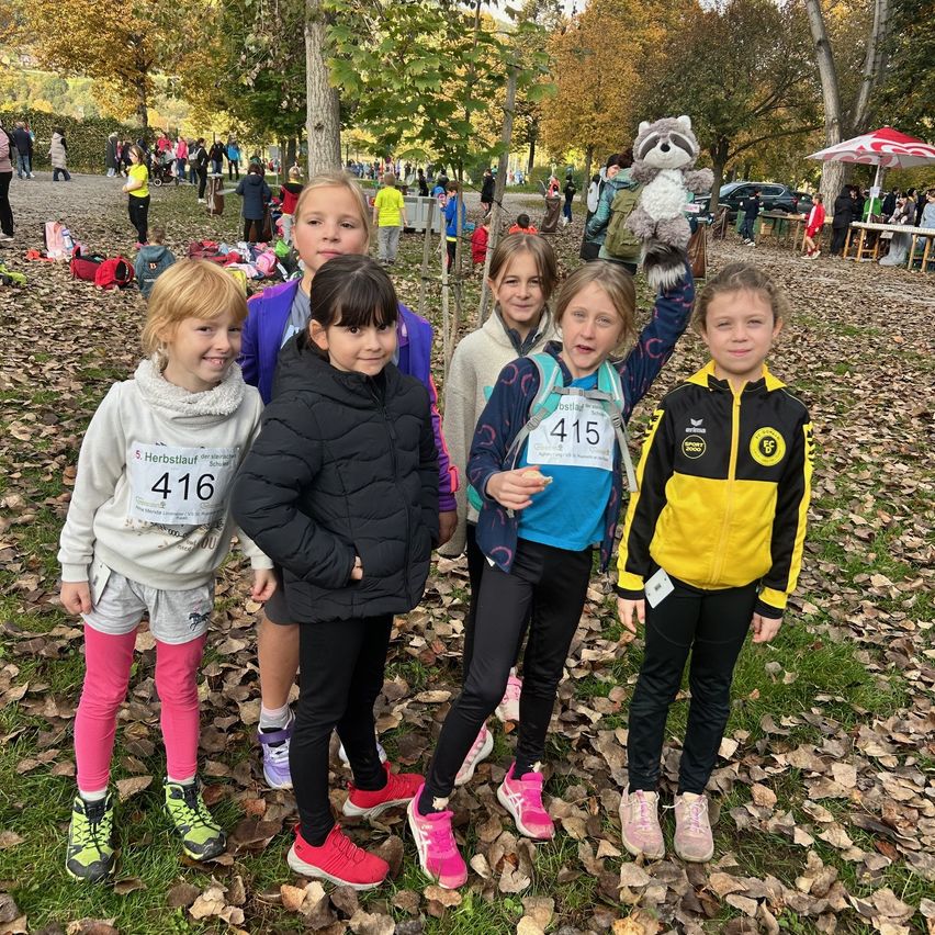 Eine Gruppe junger Mädchen steht lächelnd für ein Foto in einem Park. Sie tragen nummerierte Trikots und Turnschuhe. Eines der Mädchen hält ein Kuscheltier, während ein anderes einen Rucksack trägt. Bäume und Menschen sind im Hintergrund zu sehen.