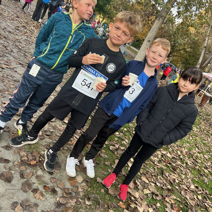 Vier Jungen stehen zusammen auf einem Pfad in einer Außenumgebung. Sie alle lächeln und scheinen für ein Foto zu posieren. Der Junge in der Mitte hält ein Sandwich und der Junge auf der rechten Seite hält einen Becher. Hinter ihnen stehen Bäume und einige Menschen.