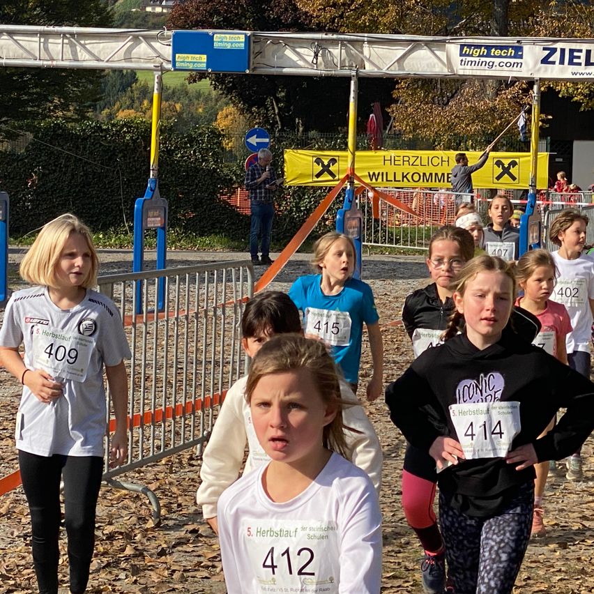 Kinder bei einem Laufrennen mit nummerierten Shirts. Ein Mann steht hinter einem Zaun mit einem Schild, das Herzlich Willkommen sagt. Bäume und Büsche sind im Hintergrund.
