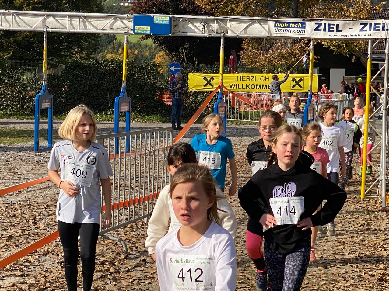 Kinder bei einem Laufrennen mit nummerierten Shirts. Ein Mann steht hinter einem Zaun mit einem Schild, das Herzlich Willkommen sagt. Bäume und Büsche sind im Hintergrund.