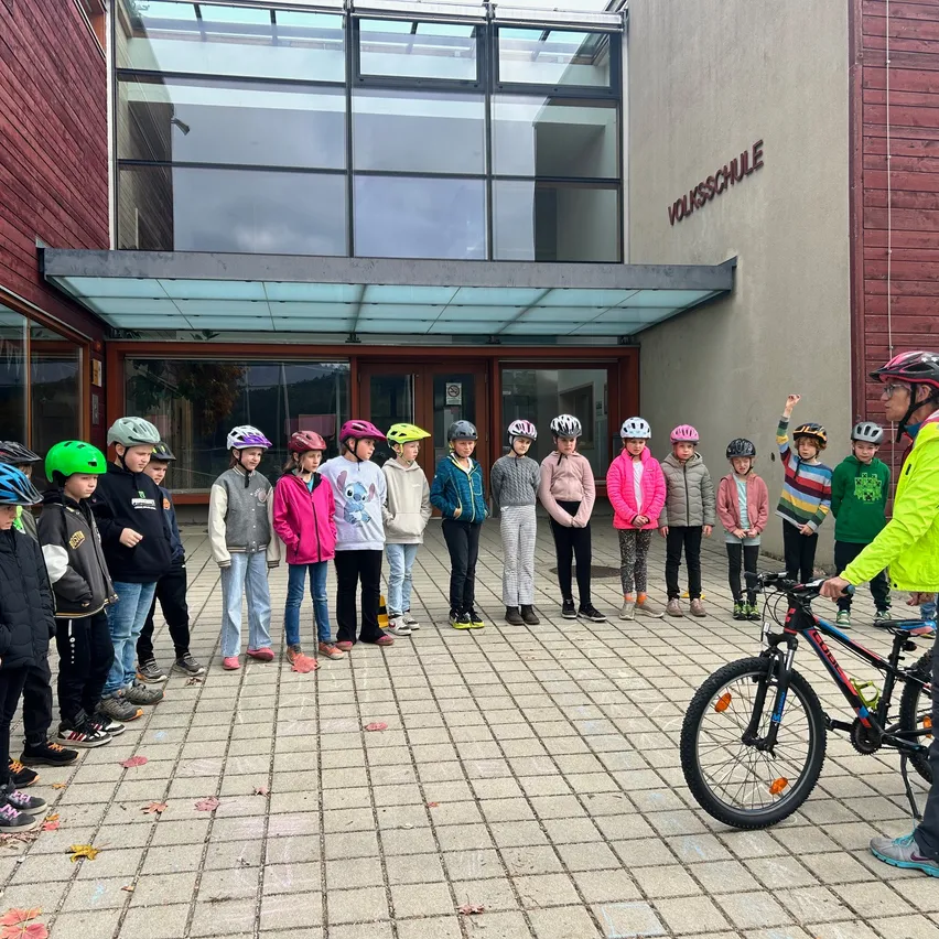 Eine Gruppe von Kindern in Helmen steht in einer Reihe vor einem Gebäude. Ein Mann mit einem Fahrrad steht vor ihnen.
