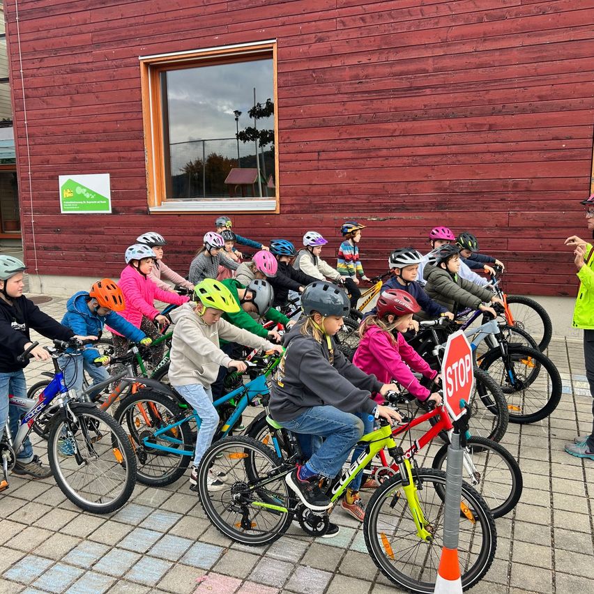 Gruppe von Kindern, die in einer Reihe Fahrräder fahren, Helme und Jacken tragen, mit einem Stoppschild und einem Gebäude im Hintergrund.