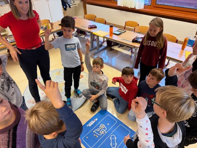 Ein Klassenzimmer mit Kindern, die sich um eine Lehrerin und ein blaues Brett versammelt haben. Ein Junge im roten Shirt kniet, andere sitzen oder stehen, und die Lehrerin trägt ein rotes Shirt.