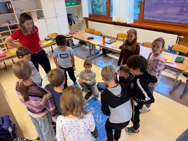 Eine Lehrerin in einem roten Shirt führt eine Gruppe von Kindern in einem Klassenzimmer an, einige stehend und andere sitzend. Die Kinder schauen auf einen blauen Teppich mit Muster auf dem Boden. Es gibt Tische und Stühle um sie herum.