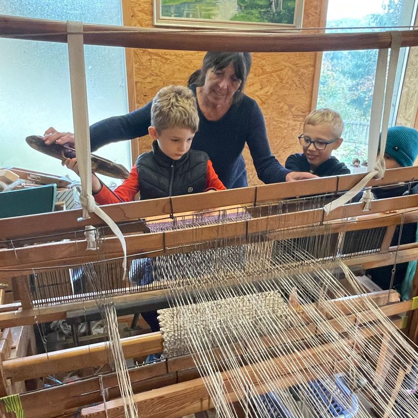 Eine Frau bringt zwei Jungen das Weben bei. Ein Junge hält ein Werkzeug, und der andere schaut sich den Stoff an. Der Webstuhl besteht aus Holz und Metall.