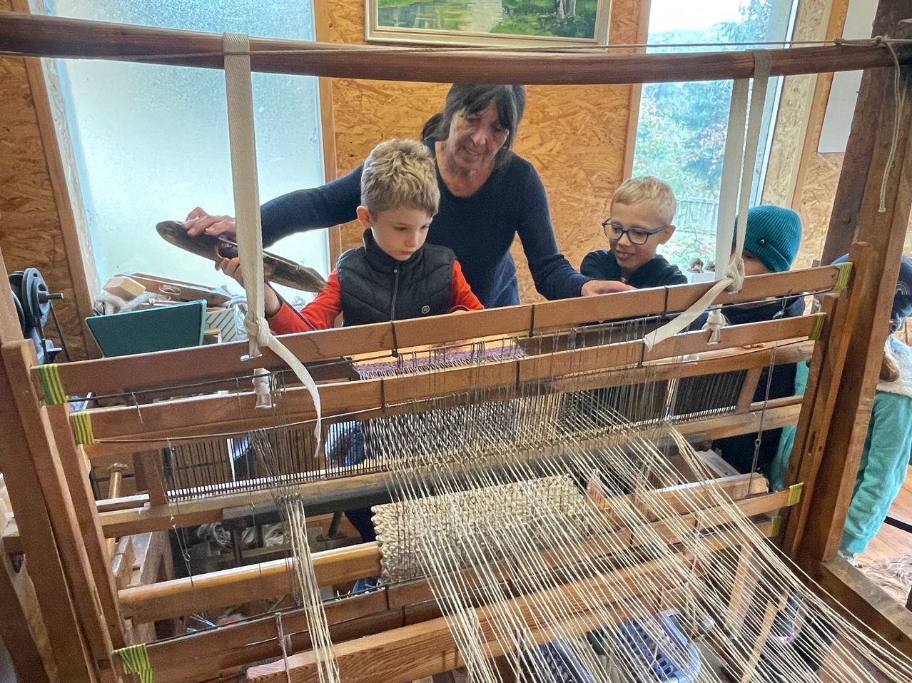 Eine Frau bringt zwei Jungen das Weben bei. Ein Junge hält ein Werkzeug, und der andere schaut sich den Stoff an. Der Webstuhl besteht aus Holz und Metall.
