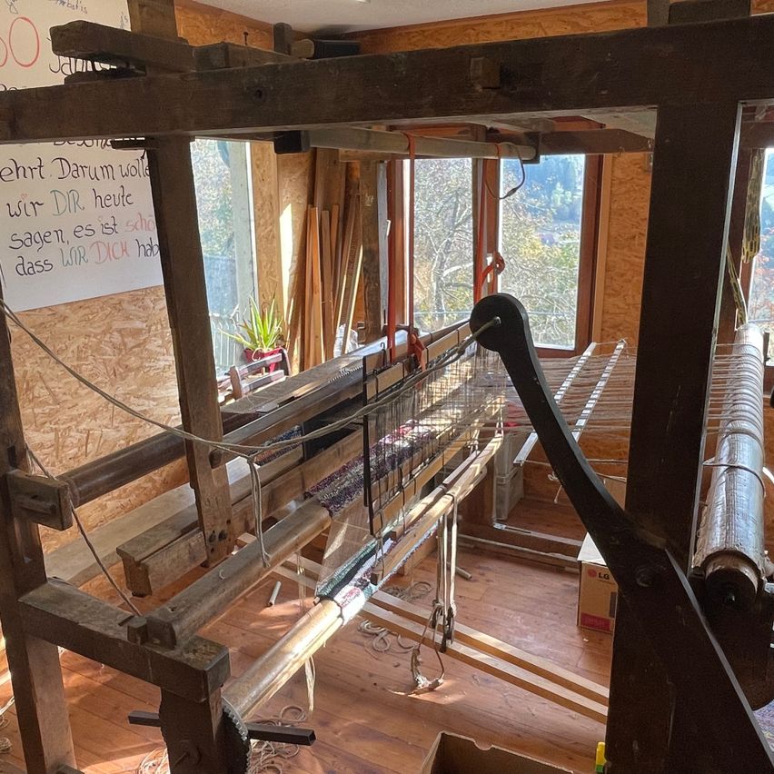 Ein Innenansicht einer Werkstatt mit einem großen Webstuhl und einer hölzernen Wand mit deutscher Schrift. Es gibt mehrere Werkzeuge und eine Topfpflanze in der Nähe der Fenster.