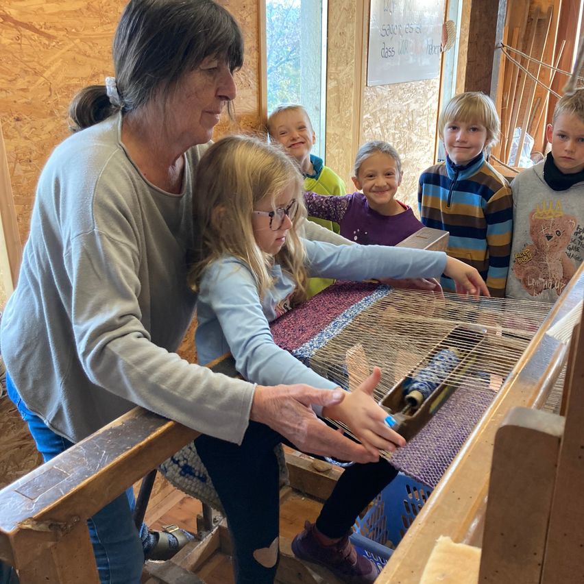 Eine ältere Frau führt ein junges Mädchen, das auf einem Webstuhl webt, umgeben von anderen Kindern. Sie befinden sich in einem Raum mit Holzwänden und einer Tafel.