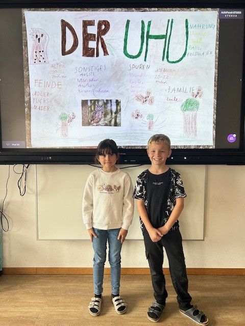 Zwei Kinder stehen vor einem Bildschirm, der ein Lehrposter über Eulen mit Text auf Deutsch und Illustrationen zeigt.
