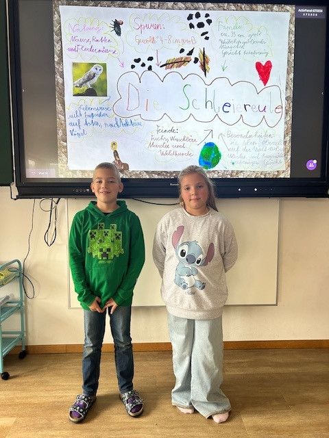 Zwei Kinder stehen vor einem großen Bildschirm, der ein Poster mit Bildungsinhalten zeigt. Sie lächeln und scheinen für ein Foto zu posieren.