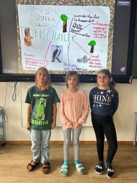 Drei junge Mädchen stehen lächelnd vor einer Tafel mit Zeichnungen. Eine von ihnen trägt ein Minecraft-Shirt. Dahinter befindet sich ein Monitor mit einer bunten Zeichnung.